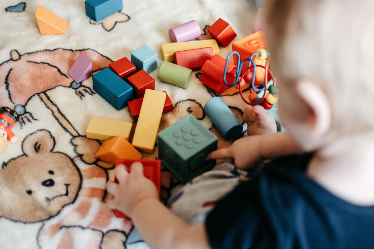 An image of a baby playing with toys. The baby is sitting on a blanket with a teddy bear holding an umbrella printed on it. The toys are wooden and plastic blocks.